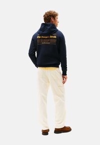 Uomo che indossa un felpa blu navy con testo stampato giallo, pantaloni cargo crema, scarpe marroni e una camicia a righe gialle, in piedi con le spalle rivolte verso la fotocamera.