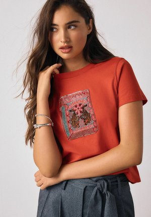 Rotes Baumwoll-T-Shirt mit dekorativem gesticktem Patch in Tiger-Design, kombiniert mit grauer, hoch taillierter Hose.