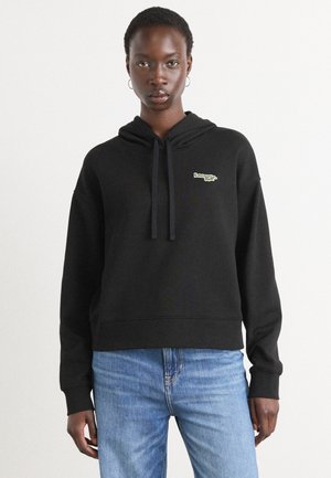 Sweatshirt à capuche noir avec un logo sur le devant en vert. Fabriqué en tissu texturé avec une coupe décontractée et des poignets en bord-côte. Associé à un jean bleu.