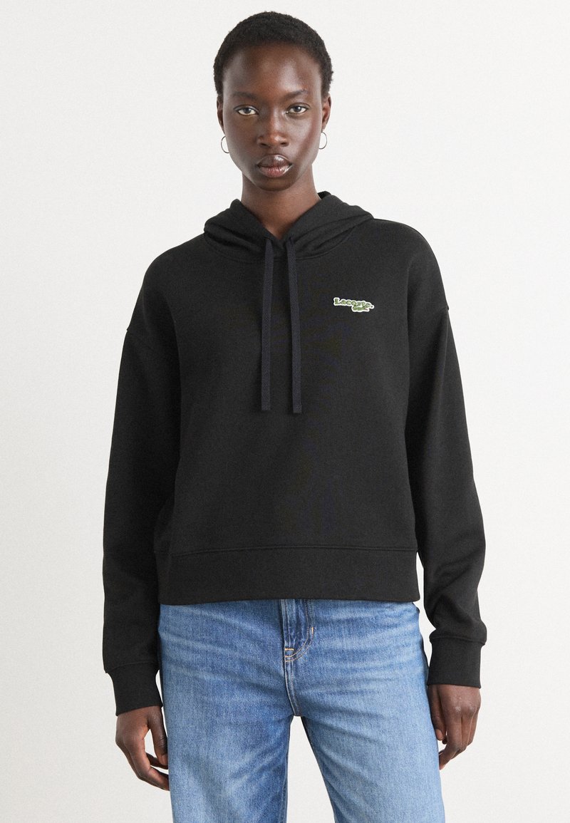 Sweatshirt à capuche noir avec un logo sur le devant en vert. Fabriqué en tissu texturé avec une coupe décontractée et des poignets en bord-côte. Associé à un jean bleu.