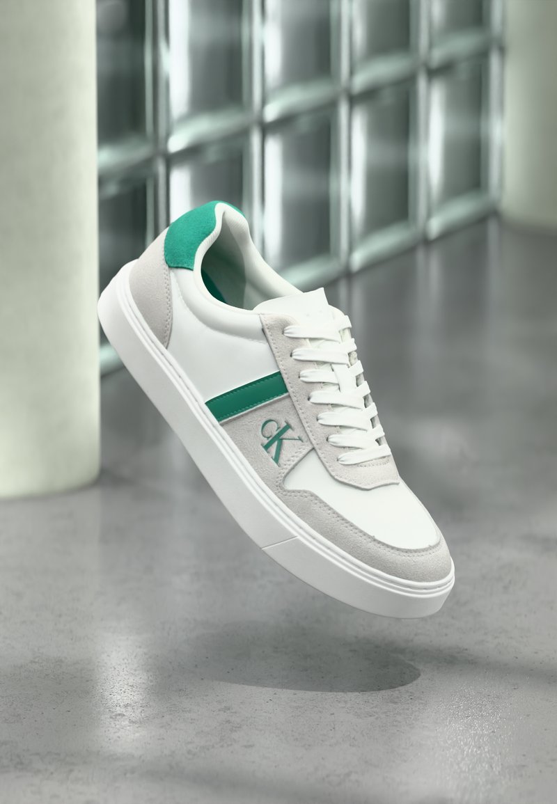 Άσπρο χαμηλό αθλητικό παπούτσι με πράσινες λεπτομέρειες, πάνελ από σουέντ, κορδόνια και λογότυπο "CK", αιωρούμενο πάνω από έναν λείο γκρι πάτωμα κοντά σε γυάλινους κύβους.