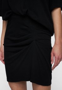 Falda negra, ajustada, hecha de tejido suave, con un diseño drapeado que presenta pliegues a los lados y un dobladillo que cae justo por encima de la rodilla.