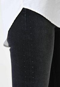 Jean en denim foncé avec une coupe ajustée, comportant de petits accents brillants le long de la couture latérale pour plus de détail. Un ourlet de chemise blanche est visible au-dessus.