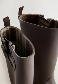 Bottes en caoutchouc jusqu'aux genoux de couleur brun foncé avec une doublure en tissu à motif tartan. Présentent un design robuste, deux languettes de tirage et un extérieur lisse.