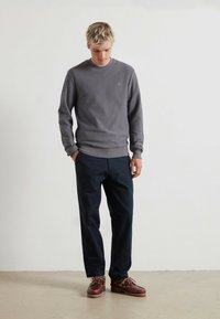 Grauer, gerippter Sweatshirt mit Rundhalsausschnitt, dunklen Hosen und braunen Bootsschuhen. Einfaches Design, strukturierter Stoff und dezentes orangefarbenes Logodetail.
