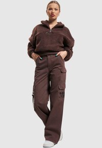 Brun fleece genser med halv glidelås og brede ermer, paret med brune cargo bukser med lommer og logo detaljer, iført hvite joggesko.