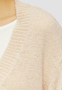 Cardigan tricoté beige clair avec un col côtelé et un design à ouvrir à l'avant. Texture douce avec une coupe ample et des détails de couture visibles.