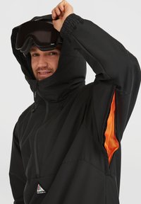 Zwarte buitenjas met capuchon, voorzien van oranje gaasaccenten, een rits aan de voorkant en elastische manchetten. Zwarte skigoggles worden boven het hoofd vastgehouden.
