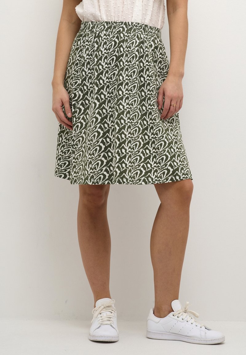 Jupe verte avec un motif abstrait blanc, taille élastique, longueur genou, fabriquée en tissu léger, associée à des baskets blanches.
