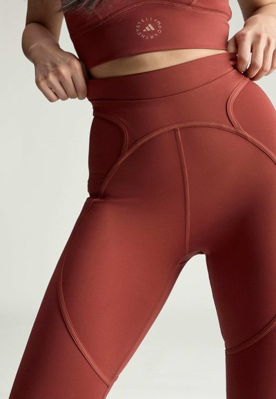 Frau trägt hoch taillierte Leggings in Rostfarbe und passendes bauchfreies Oberteil, zieht am Bund der Leggings, um die Passform anzupassen.