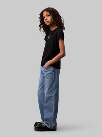 Camiseta negra de manga corta con un logo, combinada con jeans holgados de color azul claro. El modelo lleva zapatos de plataforma negros, de pie frente a un fondo gris.