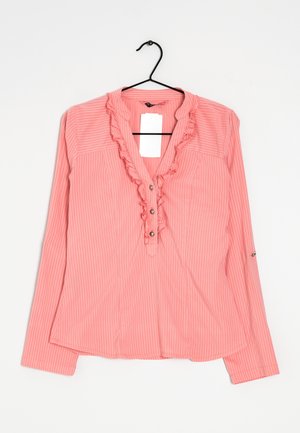 Blouse rose à manches longues avec des rayures verticales blanches, col en V volanté, boutons et une étiquette blanche, suspendue sur un cintre noir sur fond blanc.