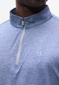 Κλείσιμο σε κοντινό πλάνο ενός ατόμου που φοράει μπλε ριγέ πουκάμισο με φερμουάρ στο ένα τέταρτο και όρθιο γιακά με λογότυπο "ck" στο στήθος.