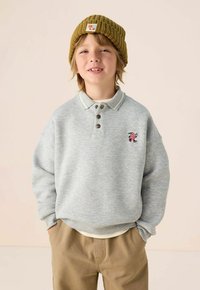 Hellgrauer Pullover-Sweatshirt mit Kragen, drei braunen Knöpfen und einem kleinen bunten gestickten Design. Getragen mit einer senfgelben Strickmütze.