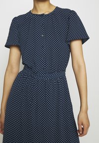Žena v námořnickomodrých šatech s bílými puntíky, s krátkým rukávem, knoflíkovým zapínáním vpředu a nařaseným pasem.