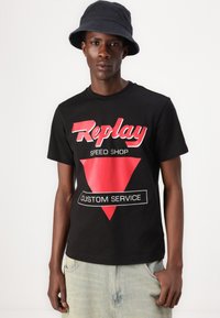 Camiseta de algodón negra con texto gráfico en rojo y blanco en un diseño triangular rojo. Combinada con shorts de mezclilla azul claro.