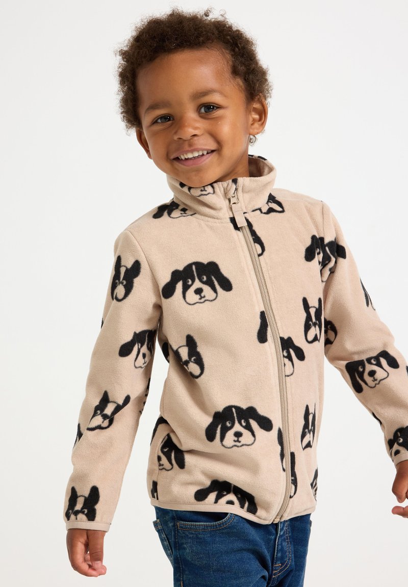 Bambino sorridente che indossa una giacca in pile beige con facce di cani neri e jeans blu, in piedi davanti a uno sfondo bianco semplice.