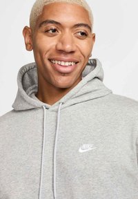 Harmaa huppari, joka on valmistettu pehmeästä materiaalista, ja siinä on etutasku sekä valkoinen Nike-logo vasemmassa rinnassa. Hupparissa on nauhat ja se on suunniteltu rentoon istuvuuteen.