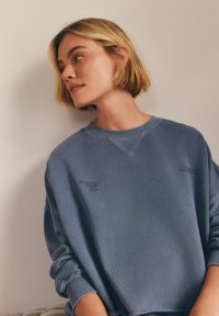 Niebieski bawełniany sweatshirt z prążkowanymi mankietami i dekoltem. Posiada haftowany napis na klatce piersiowej, luźny krój oraz teksturowany materiał.