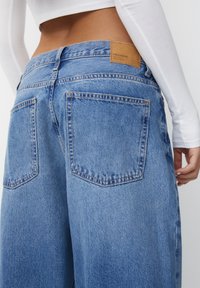 Jeans de mezclilla azul claro con cintura alta, pierna ancha y bolsillos traseros. Presenta una etiqueta de marca de cuero marrón claro en la cintura.