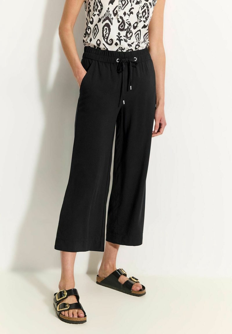 Personne portant un pantalon noir large et court à la taille élastique, des sandales noires à boucle, et un haut sans manches à motifs