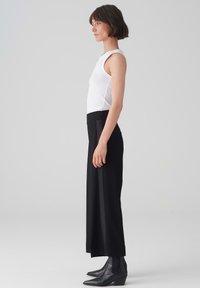 Pantalons larges noirs avec une texture lisse et une taille haute, associés à un débardeur blanc ajusté, vus de côté. Tissu léger.