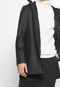 Schwarze wasserdichte Jacke mit Reißverschluss und Druckknopfverschluss, ausgestattet mit einer Kapuze, zwei Fronttaschen und einer glatten, matten Oberfläche.