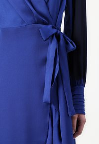 Μπλε φόρεμα με υφή, λεπτομέρεια στη μέση με κόμπο και μακριά μανίκια με πτυχωτές λεπτομέρειες. Το φόρεμα είναι κυρίως σε βασιλικό μπλε χρώμα.