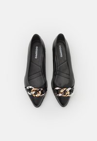 Zapatos planos negros con un acabado de cuero suave. Presentan un detalle decorativo en cadena en oro, plata y negro. El interior es acolchado y lleva la marca.