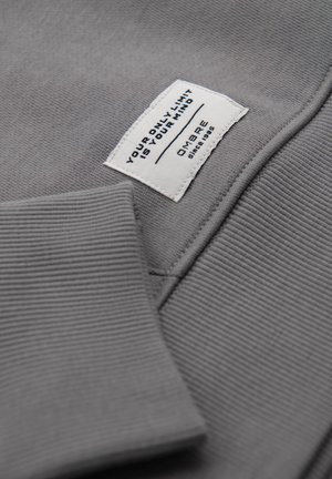 Tessuto di felpa grigia con polsini a coste, con etichetta bianca con testo nero che dice, "IL TUO UNICO LIMITE È LA TUA MENTE," e nome del marchio, OMBRE.