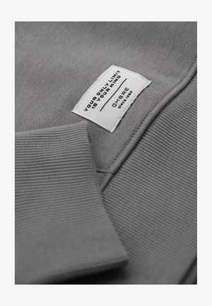 Harmaa collegepaita, jossa on ribbireunat, ja jossa on valkoinen etiketti mustalla tekstillä, joka lukee: "AINOA RAJA ON MIELESI," sekä brändäys.