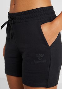 Mustat urheilusortsit, jotka on valmistettu pehmeästä jersey-kankaasta, jossa on joustava vyötärö, naruhäntäyksityiskohtia ja sivutaskut. Lahkeissa on logoprintti.