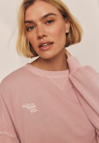 Růžová bavlněná mikina s kulatým výstřihem a spadlými rameny. Na levé části hrudi je bíly nápis "Wellness team". Měkká textura.