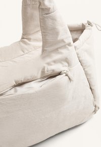 Beige puffy taske med to håndtag, der har en lynlås åbning og en glat stofstruktur. Rund form med syet detaljer.