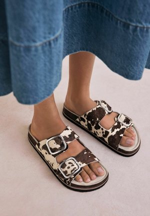 Füße mit Sandalen im Kuhmuster mit zwei silbernen Schnallen, kombiniert mit einem blauen Jeansrock, stehen auf einem hellen Boden.