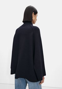 Strikket marineblå kappe med høy krage, vide ermer og avslappet passform. Glatt stoffstruktur. Kombinert med lyseblå slitte jeans.