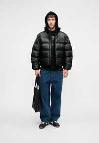 Homem vestido com uma jaqueta acolchoada preta e capuz, jeans azuis largos, sapatilhas pretas, segurando uma bolsa tote preta, de pé contra um fundo branco.