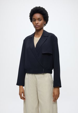 Kvinde iført en sort cropped blazer over en beige top og lyse beige løstsiddende bukser, stående mod en hvid baggrund.