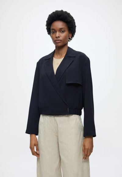 Femme portant un blazer court noir sur un top beige et un pantalon ample beige clair, debout sur un fond blanc.