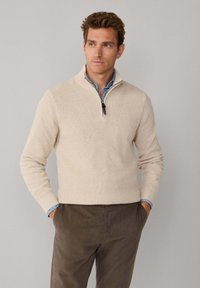 Beige pullovertrui met een halve rits hals, geribbelde textuur, lange mouwen en een aansluitend ontwerp, gecombineerd met een lichtblauw overhemd met kraag.