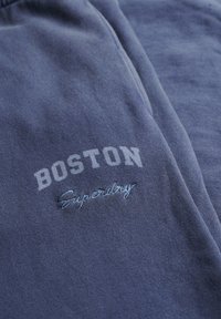 Blå sweatshirtstof med 'BOSTON' trykt i hvid og 'Superdry' broderet i lys blå. Blød tekstur og afslappet design.