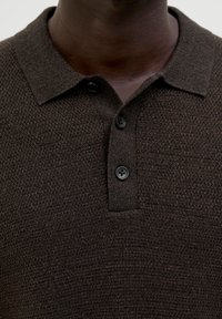 Braunes strukturiertes Poloshirt mit klassischem Kragen, ausgestattet mit drei schwarzen Knöpfen an der Knopfleiste. Der Stoff wirkt weich und gestrickt.