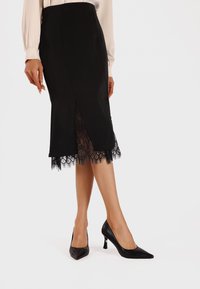 Gonna nera a matita in tessuto liscio con orlo in pizzo, dotata di spacco frontale e abbinata a décolleté nere con punta a punta.