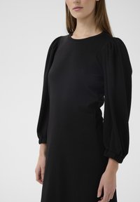 Rochie neagră cu decolteu rotund și mâneci lungi bufante. Confecționată dintr-un material neted, cu croială lejeră și manșete adunate.