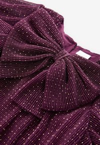 Gros plan sur un tissu plissé violet avec des pois dorés et un grand nœud décoratif au niveau de l'encolure d'un vêtement.