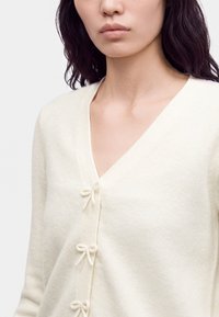 Femme portant un cardigan crème à encolure en V avec trois petites fermetures en nœud sur le devant et des cheveux ondulés foncés sur les épaules.