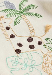 Geborduurde stof met een giraffe met bruine vlekken, palmbomen en tropische bladeren in groen en lichtblauw. De textuur is zacht en gestructureerd.