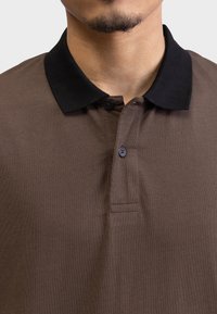 Brown polo shirt featuring a black ribbed collar, two dark buttons, and a smooth texture. Close-up view focused on neckline and chest area.