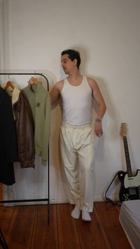 Homme en débardeur blanc et pantalon choisissant une veste verte dans un portant à vêtements, dans une pièce avec un sol en bois et une guitare électrique à proximité.