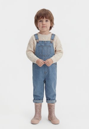 Bambino piccolo in piedi con maglione crema, salopette di jeans risvoltate e stivali da pioggia rosa chiaro con motivo su sfondo bianco.